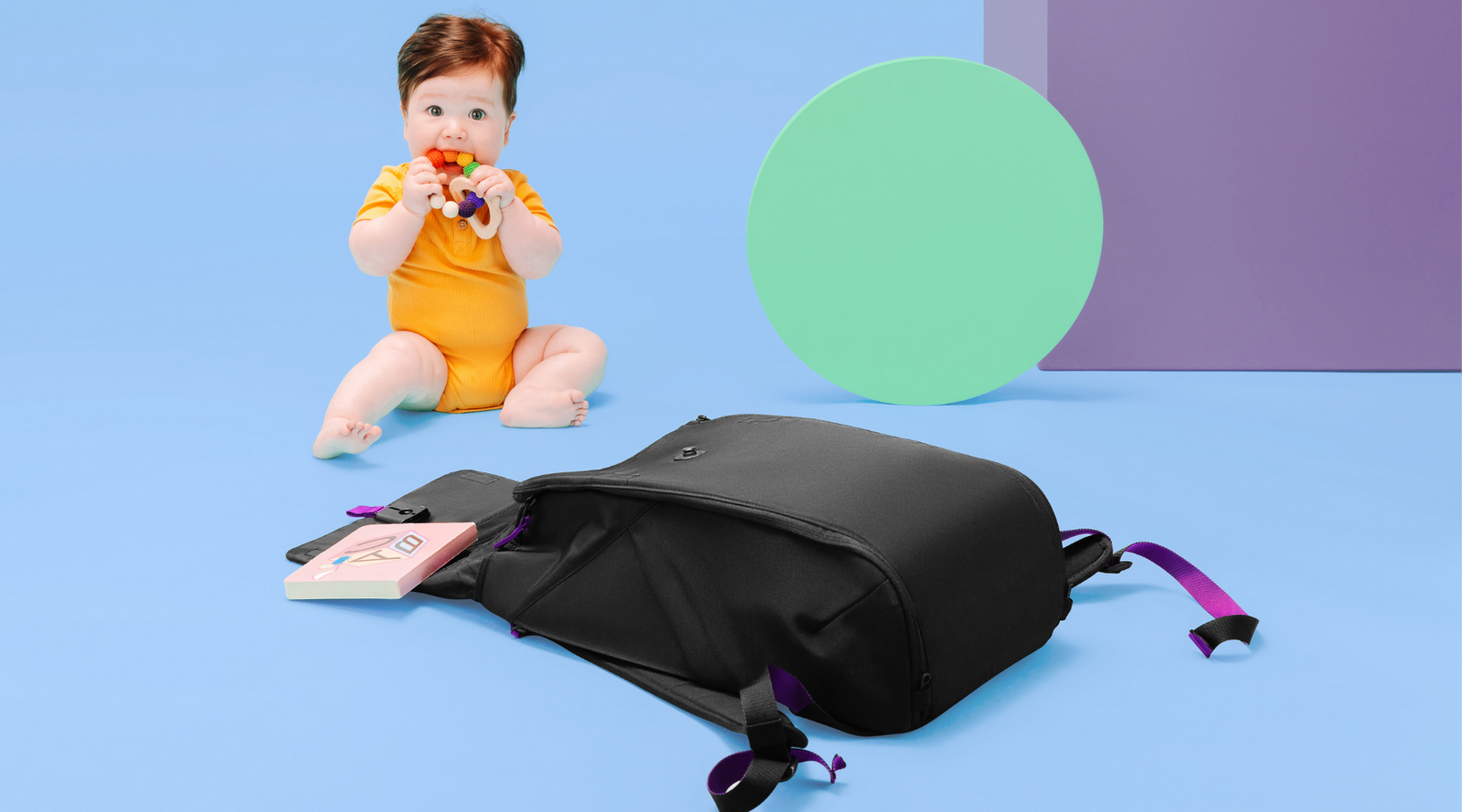 Black backpack laying open on blue background. Baby in orange sitting in background next to teal and purple decorative shapes.