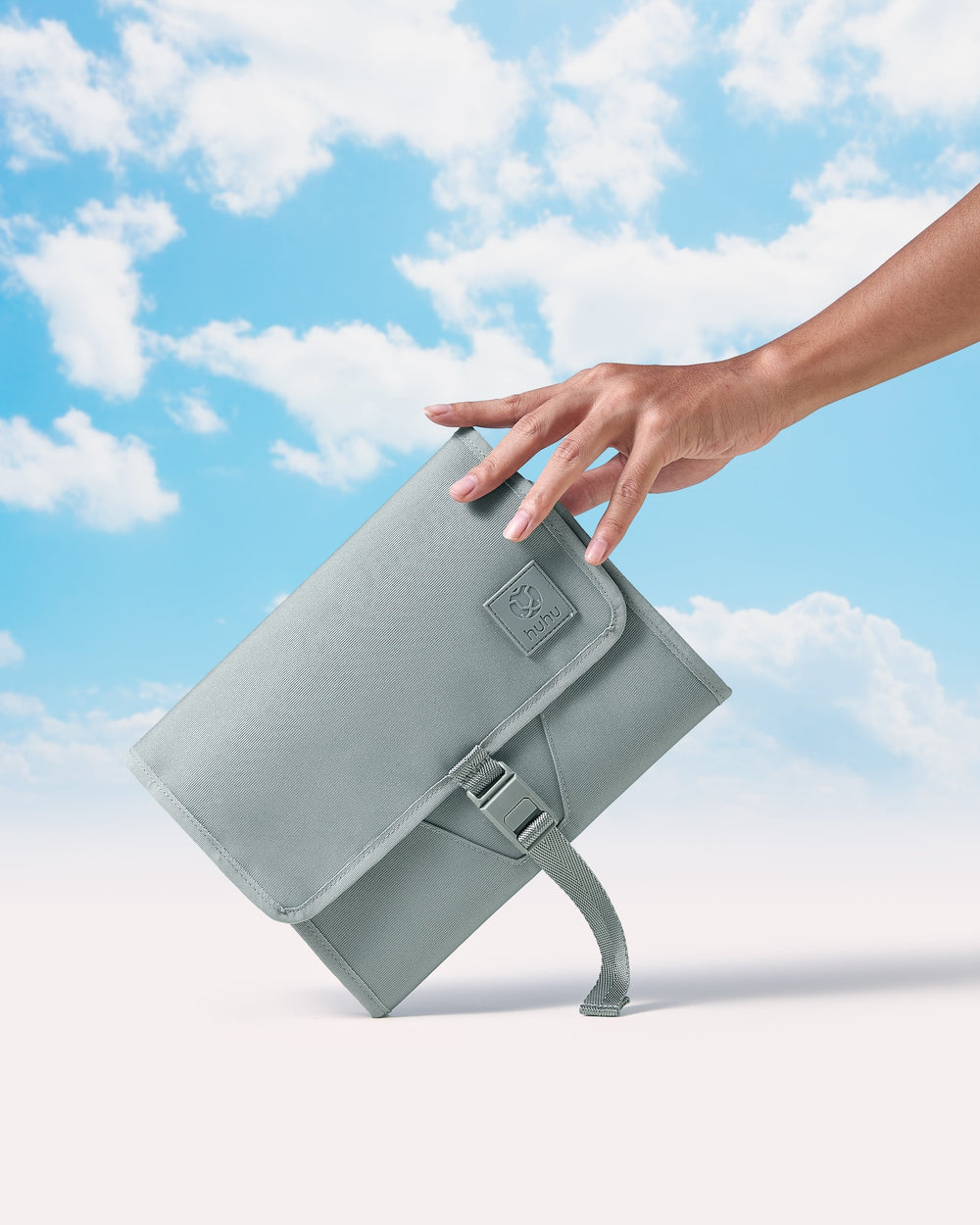 Hand holding green changing wallet on angle against beige and blue background with white clouds.