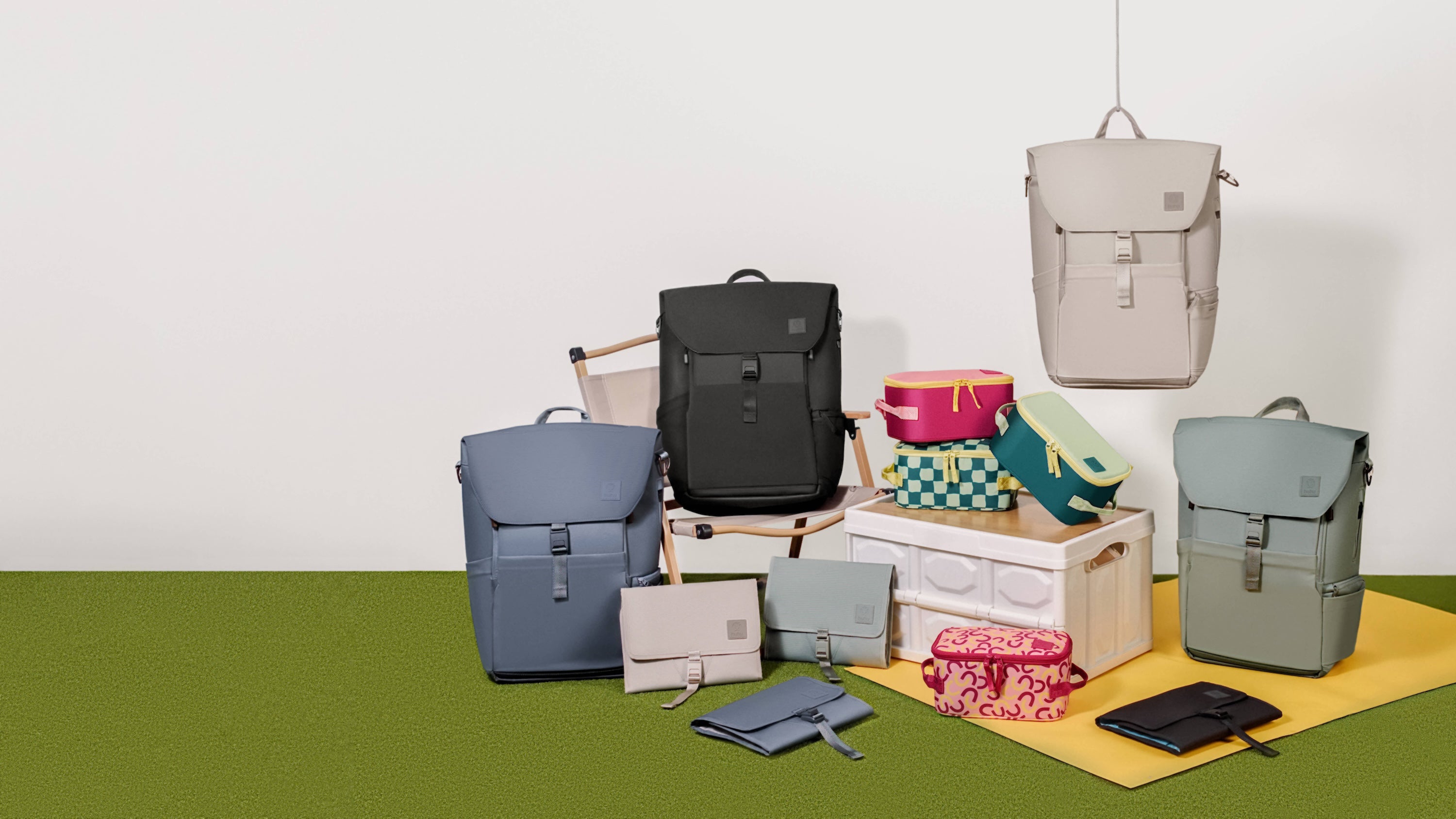 Collection of backpacks, changing wallets, and insulated cooler bags. All colors in collection sitting atop chair, box, and yellow blanket. White background with green ground.