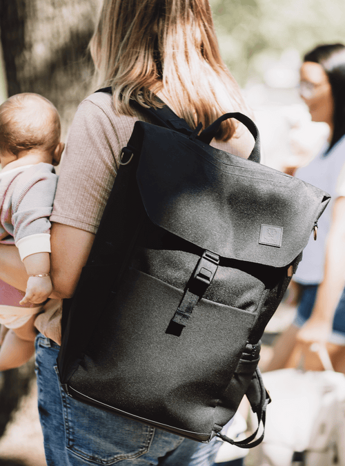 Mom holding a baby faced away from the camera. She is wearing a black backpack. Background is blurred view of another mom.