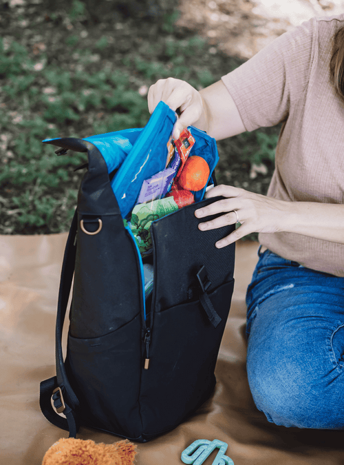 Mom wearing mauve shirt and blue jeans sitting on a brown blanket in the grass. Her hands are reaching into an opened up black backpack with a blue interior. She is pulling out a blue packing pouch filled with snacks.