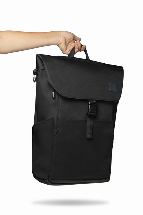Hand holding black backpack by its top handle against white background.