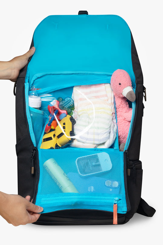 Front view of hands opening a black backpack to reveal blue interior with blue packing pouch. Backpack and packing pouch are packed with brightly colored children's items. White background.