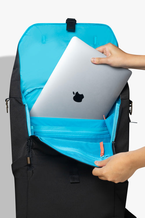 Front view of hands placing silver laptop into padded laptop pocket of an opened black backpack against a white background. Interior of bag is blue, with an orange zipper pull showing.