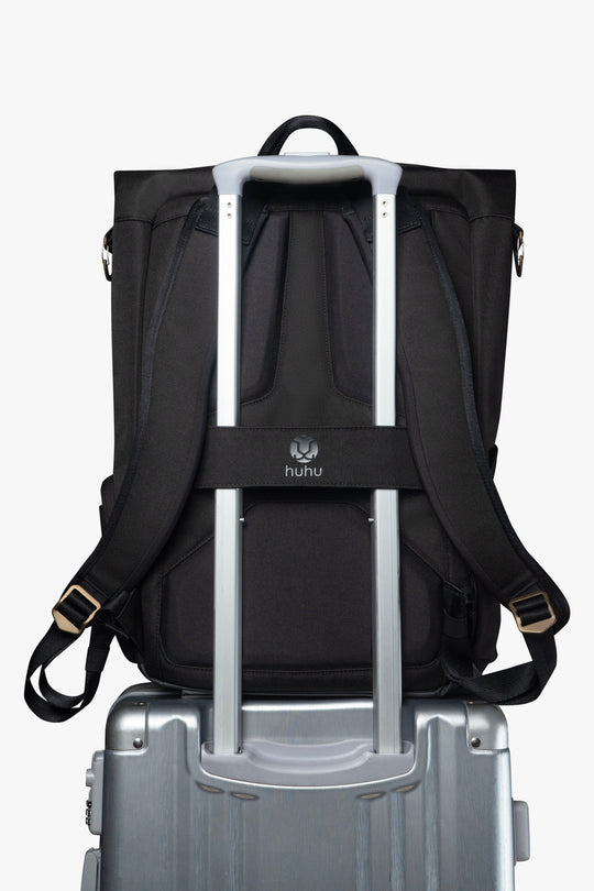 Back view of black backpack. Attached luggage strap is holding backpack to the handle of a grey rolling suitcase.