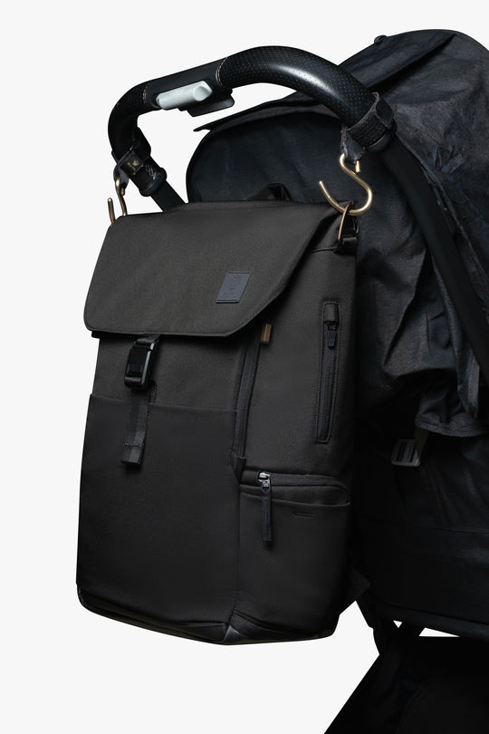 Black backpack hanging by gold stroller hooks on handle of black stroller against a white background.