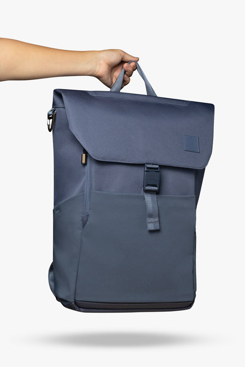 Hand holding blue backpack by its top handle against white background.