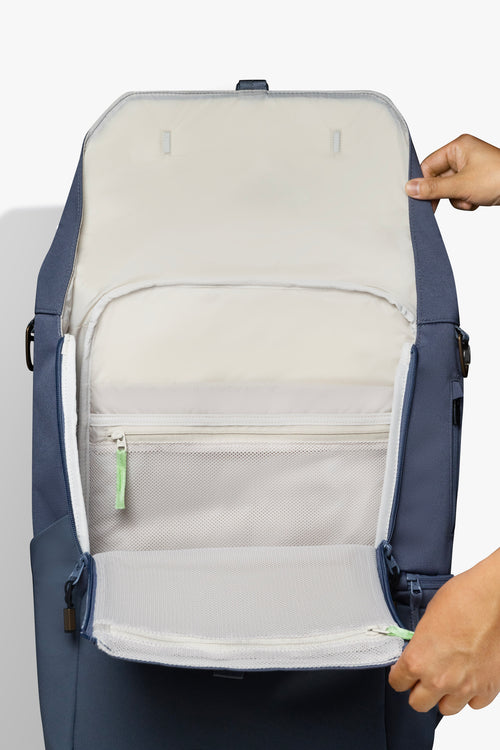Front view of hands holding an opened blue backpack against white background. Image shows taupe interior of backpack and green zipper pull.