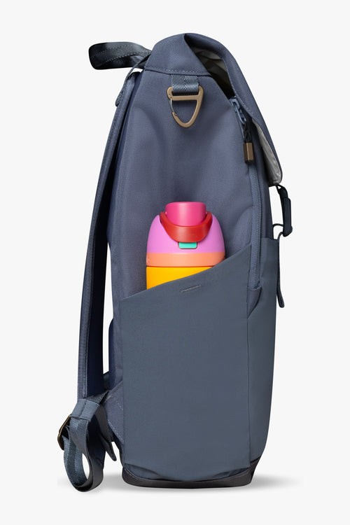Side view of blue backpack against a white background. Showing stretchy bottle pocket on side of backpack holding brightly colored yellow and pink water bottle. Bronze carabiner clip and blue backpack straps are visible.
