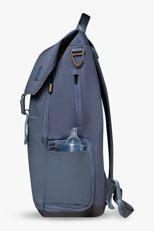 Side view of blue backpack against a white background. Showing baby bottle in side stretchy bottle pocket. Bronze bag hardware and exterior zippered white pocket are visible.
