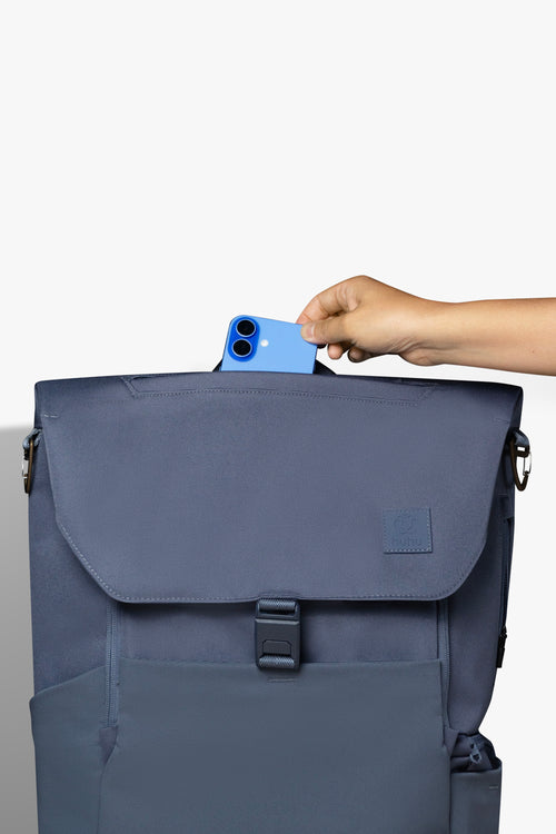 Front view of blue backpack against white background. Hand is placing blue phone into zipper pocket on top of backpack.