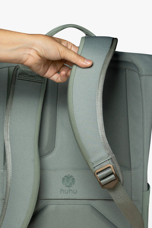 Back view of green backpack against white background. Hand is touching padded strap of backpack.