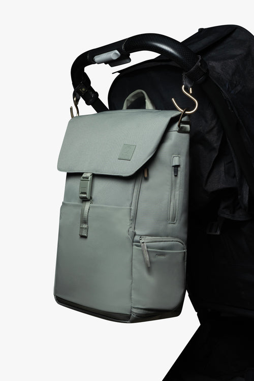 Green backpack hanging by gold stroller hooks on handle of black stroller against a white background.