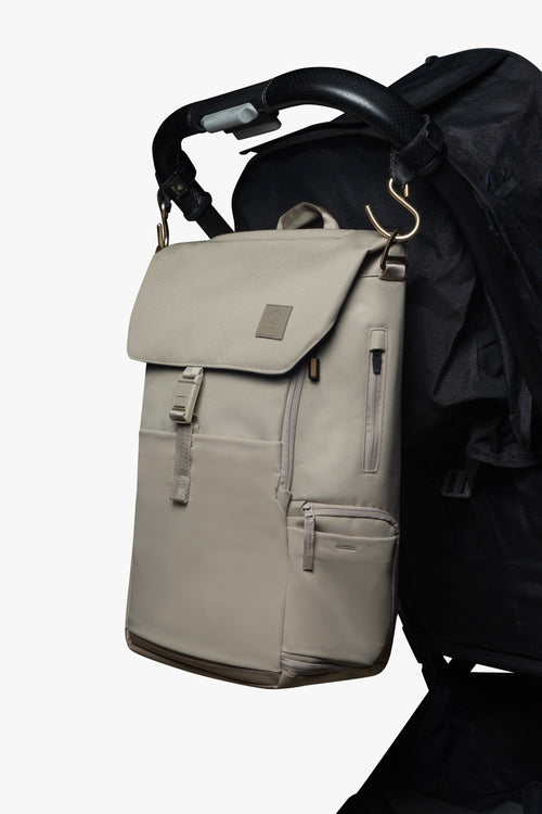 Taupe backpack hanging by gold stroller hooks on handle of black stroller against a white background.