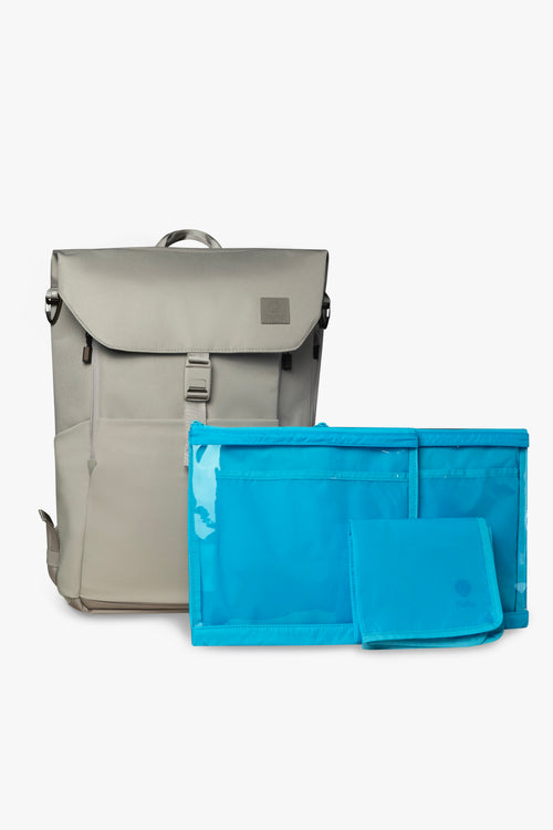 Front view of taupe backpack, closed and standing upright with 2 blue packing pouches and 1 blue folded changing pad against white background.
