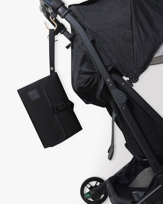 Black changing wallet with strap hanging from stroller hook on a black stroller against a white background.