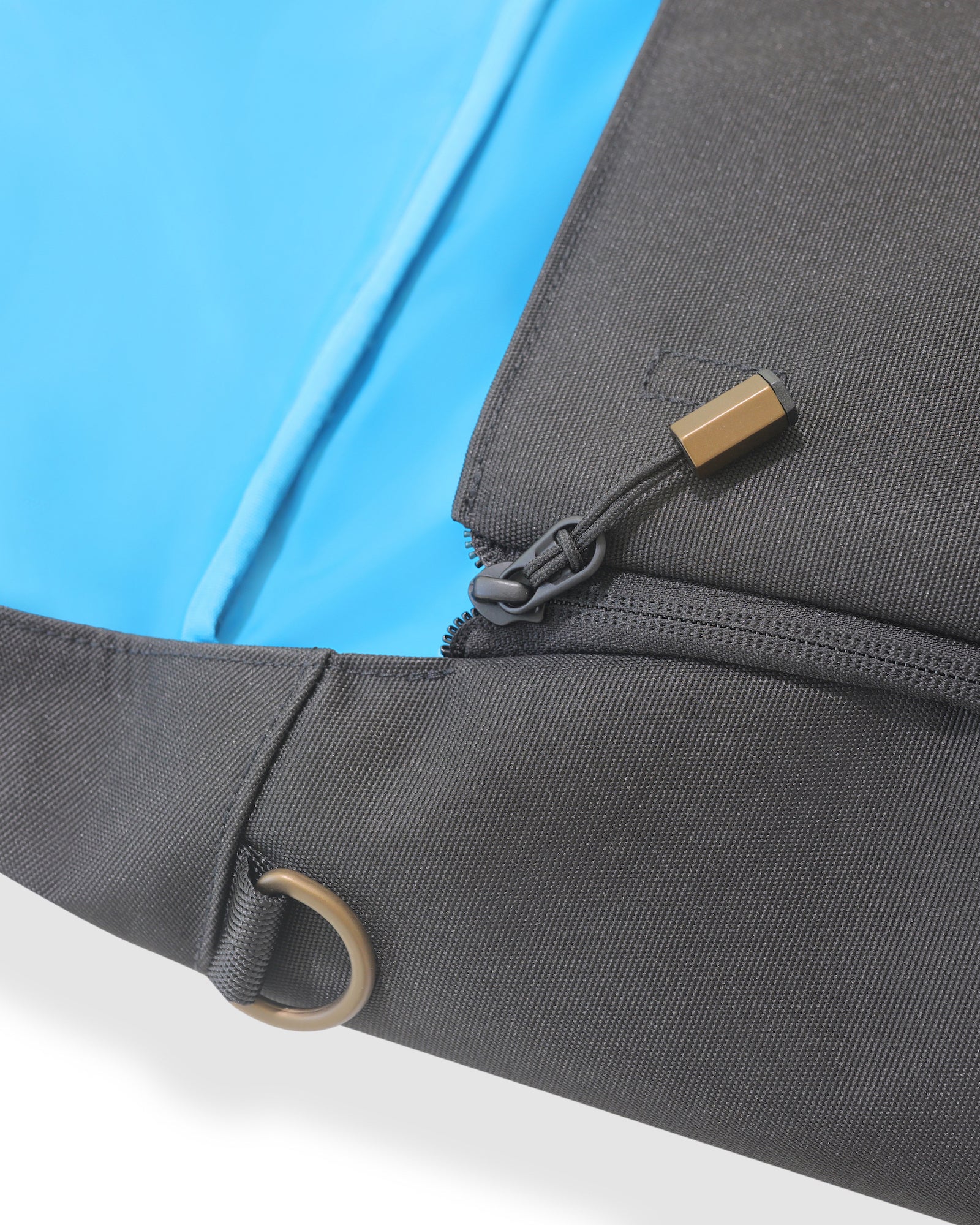 Close up of gold and black hardware on black backpack against a white background. Image zoomed in on black zipper and gold hook, and blue interior of backpack.