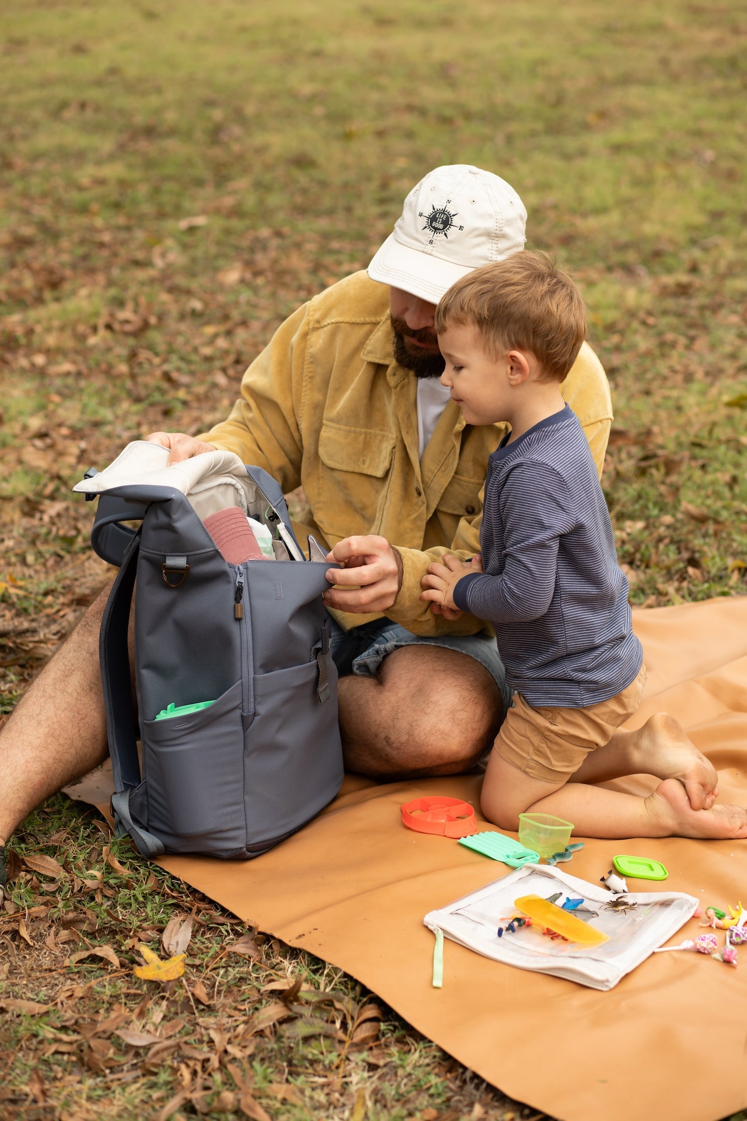 Man in white hat and yellow shirt sitting with a young boy wearing a blue shirt on a picnic blanket in a park. Man is opening a diaper bag for the son to look into.