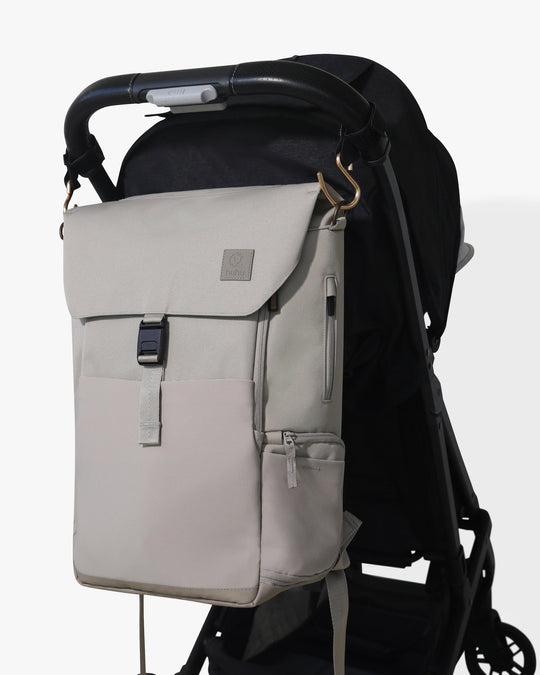 Taupe backpack with hooks hanging from stroller hooks on a black stroller against a white background.