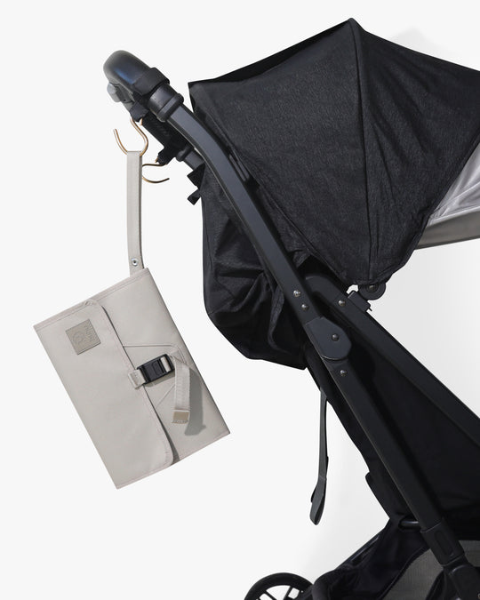 Taupe changing wallet with strap hanging from stroller hook on a black stroller against a white background.
