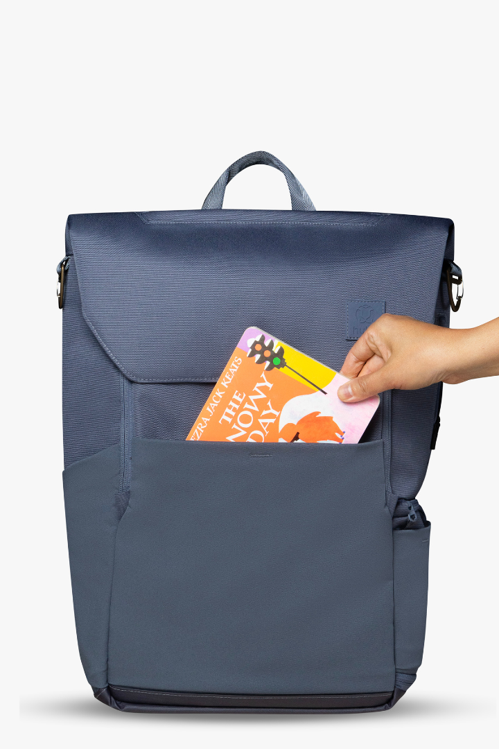 Front view of blue backpack with hand placing orange and yellow board book into front stretchy pocket of backpack.