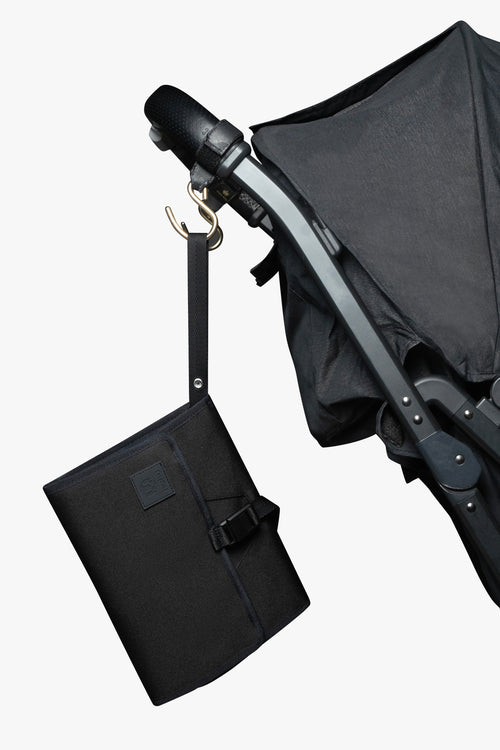 Black changing wallet hanging from strap attached to gold stroller hook on a black stroller against white background.