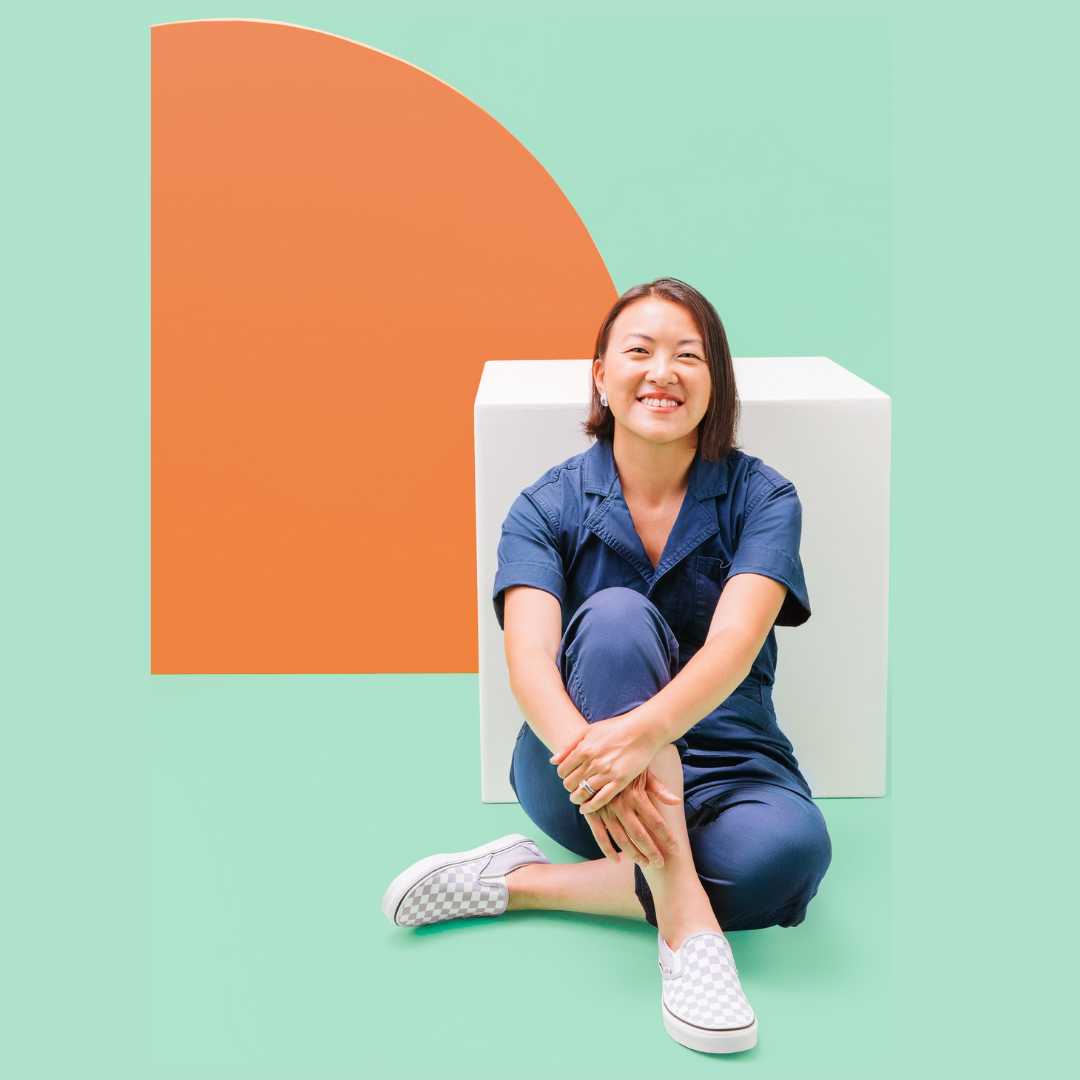 Meng, founder of Huhu, sitting and smiling with legs crossed in a teal room with white and orange decorative shapes.