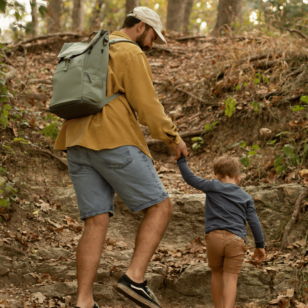 Dad in yellow shirt wearing green backpack, holding hands with toddler as they hike up a trail in a wooded area.