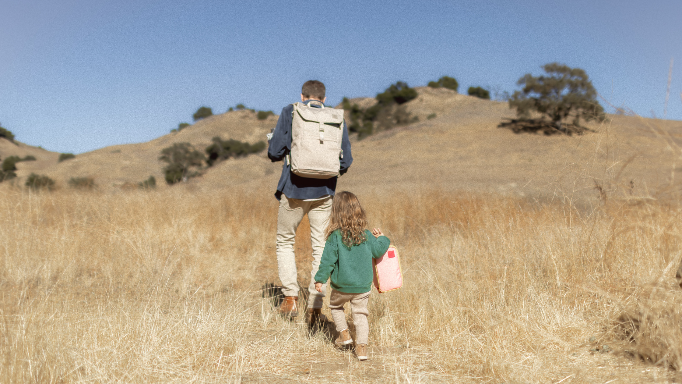 Dad wearing diaper backpack and daughter holding cooler bag walking outdoors. Grassy hill and sky in background.