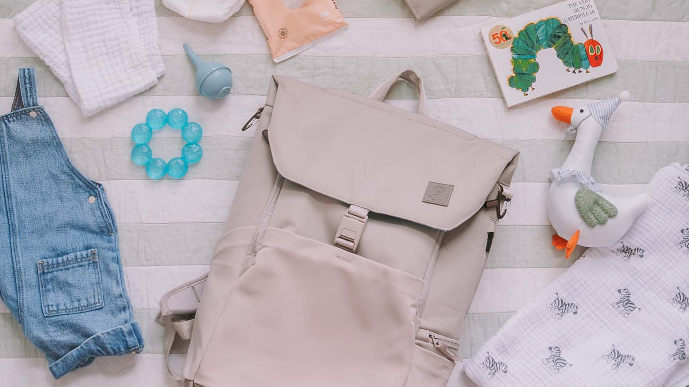 Flat lay of baby items on a green and white striped blanket. Beige diaper backpack at center of image.