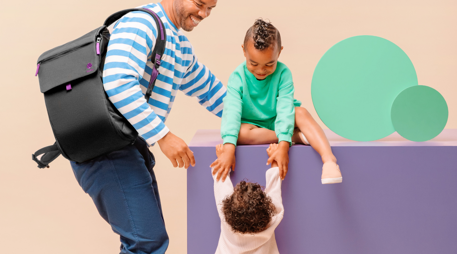 Dad wearing black backpack interacting with two kids. One child sitting on purple box, helping smaller child up. Green decorative circles against a beige background.