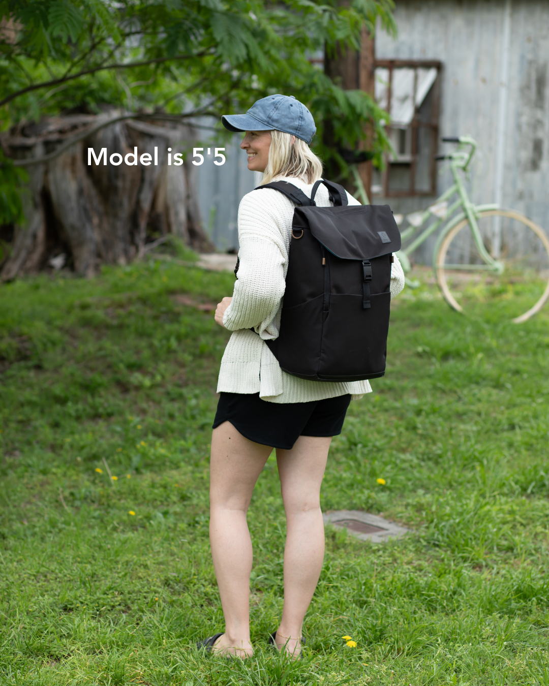 Women wearing tan sweatshirt, blue hat, and black shorts with a black backpack standing in grass. Bicycle and trees in background. Words reading 'Model is 5'5' on left side of image.