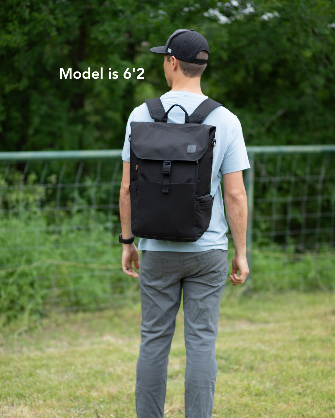 View from behind of a man wearing black hat, a blue shirt, and grey pants with a black backpack on his back. The background is grass and trees, with a green fence and 'Model is 6'2 ' written in white on left side.