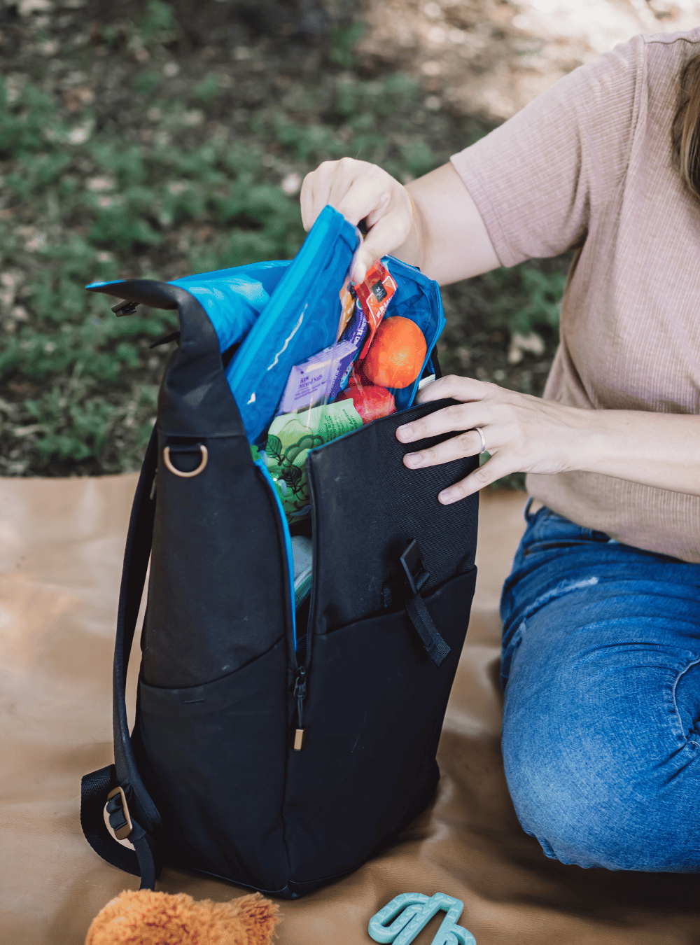 Mom wearing mauve shirt and blue jeans sitting on a brown blanket in the grass. Her hands are reaching into an opened up black backpack with a blue interior. She is pulling out a blue packing pouch filled with snacks.