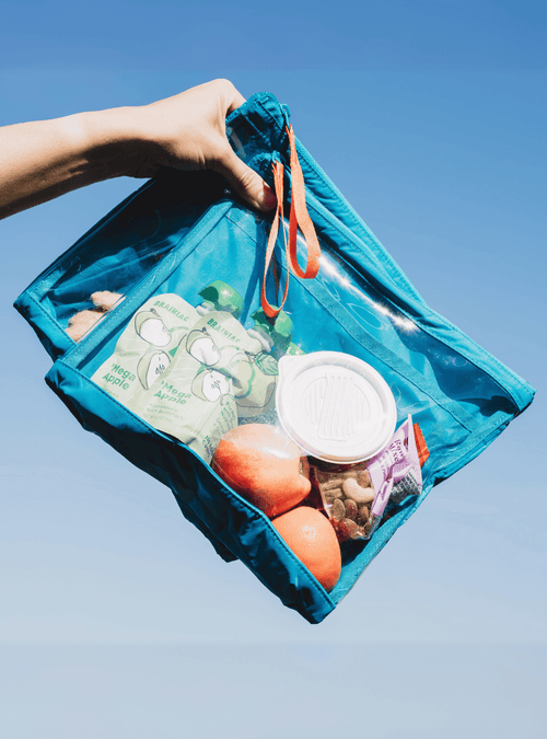 Hand holding two blue packing pouches with orange pull strings against a blue sky background. Pouches have a clear front, showing snacks packed inside.