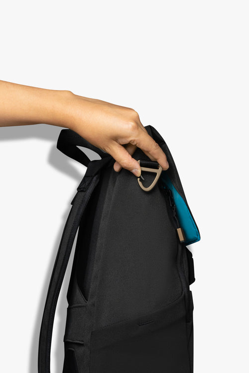 Hand opening bronze carabiner clip on side of black backpack against a white background. Blue interior of bag peaks out on top flap of backpack.