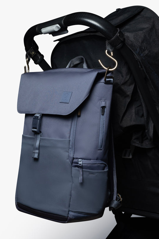 Blue backpack hanging by gold stroller hooks on handle of black stroller against a white background.