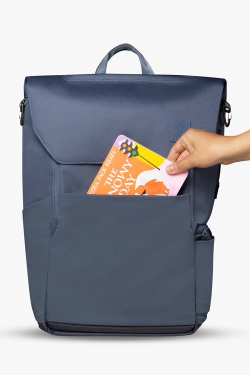 Front view of blue backpack against white background. Hand is placing orange and yellow board book into front stretchy pocket of backpack.
