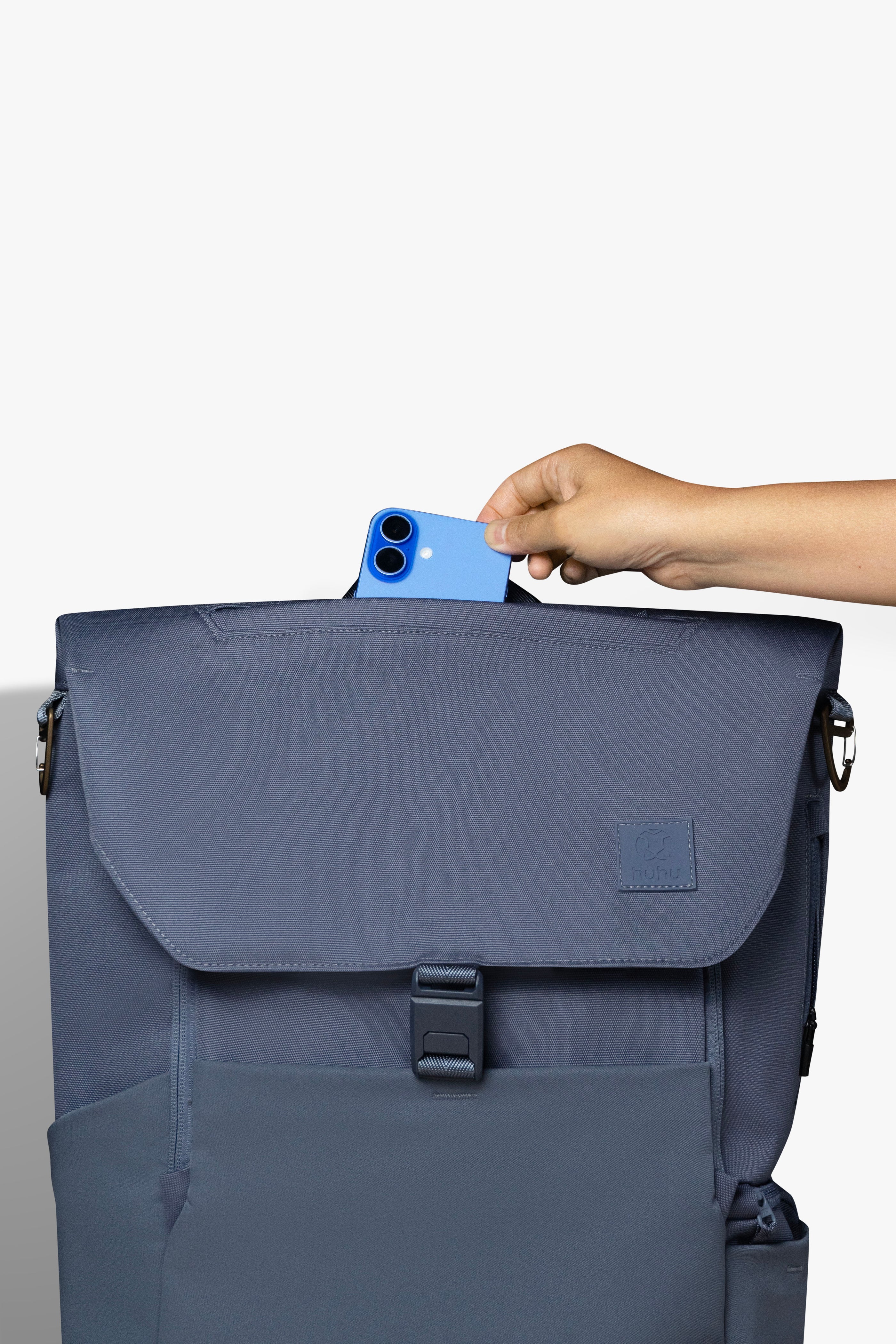 Front view of blue backpack against white background. Hand is placing blue phone into zipper pocket on top of backpack.