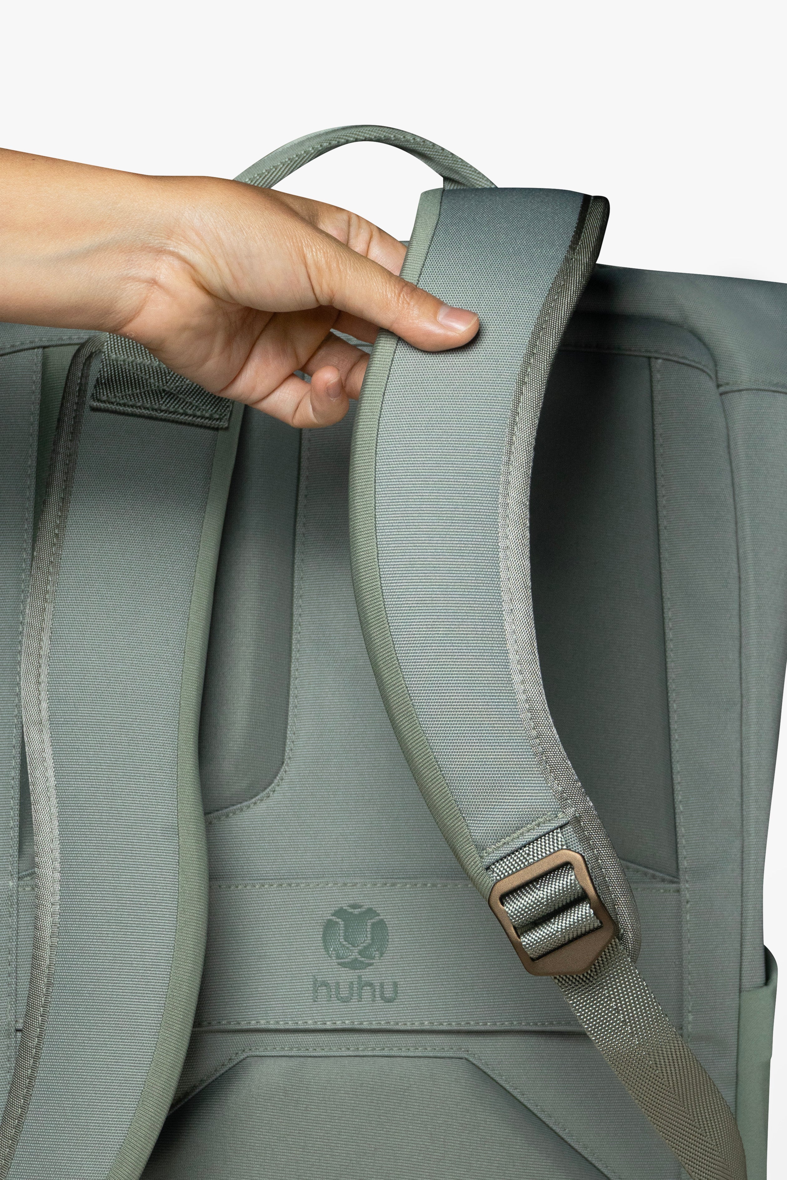 Back view of green backpack against white background. Hand is touching padded strap of backpack.