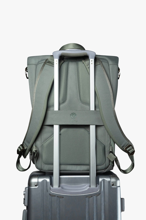 Back view of green backpack. Attached luggage strap is holding backpack to the handle of a grey rolling suitcase.