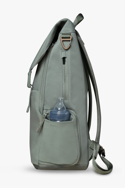 Side view of green backpack against a white background. Showing baby bottle in side stretchy bottle pocket. Bronze bag hardware and exterior zippered wipes pocket are visible.