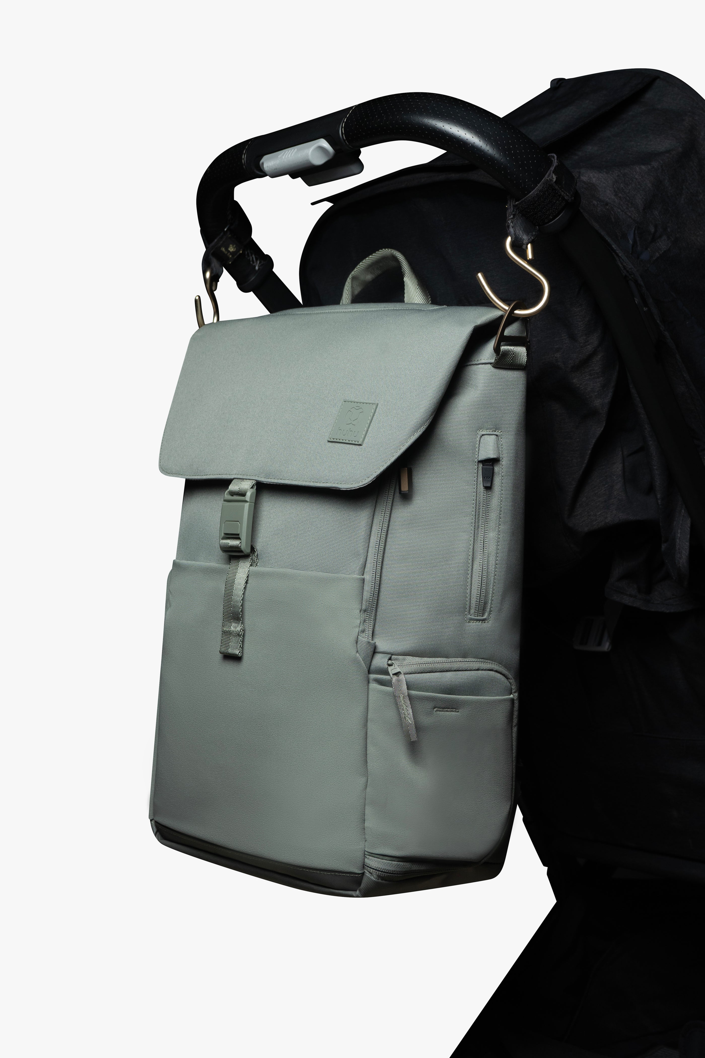 Green backpack hanging by gold stroller hooks on handle of black stroller against a white background.