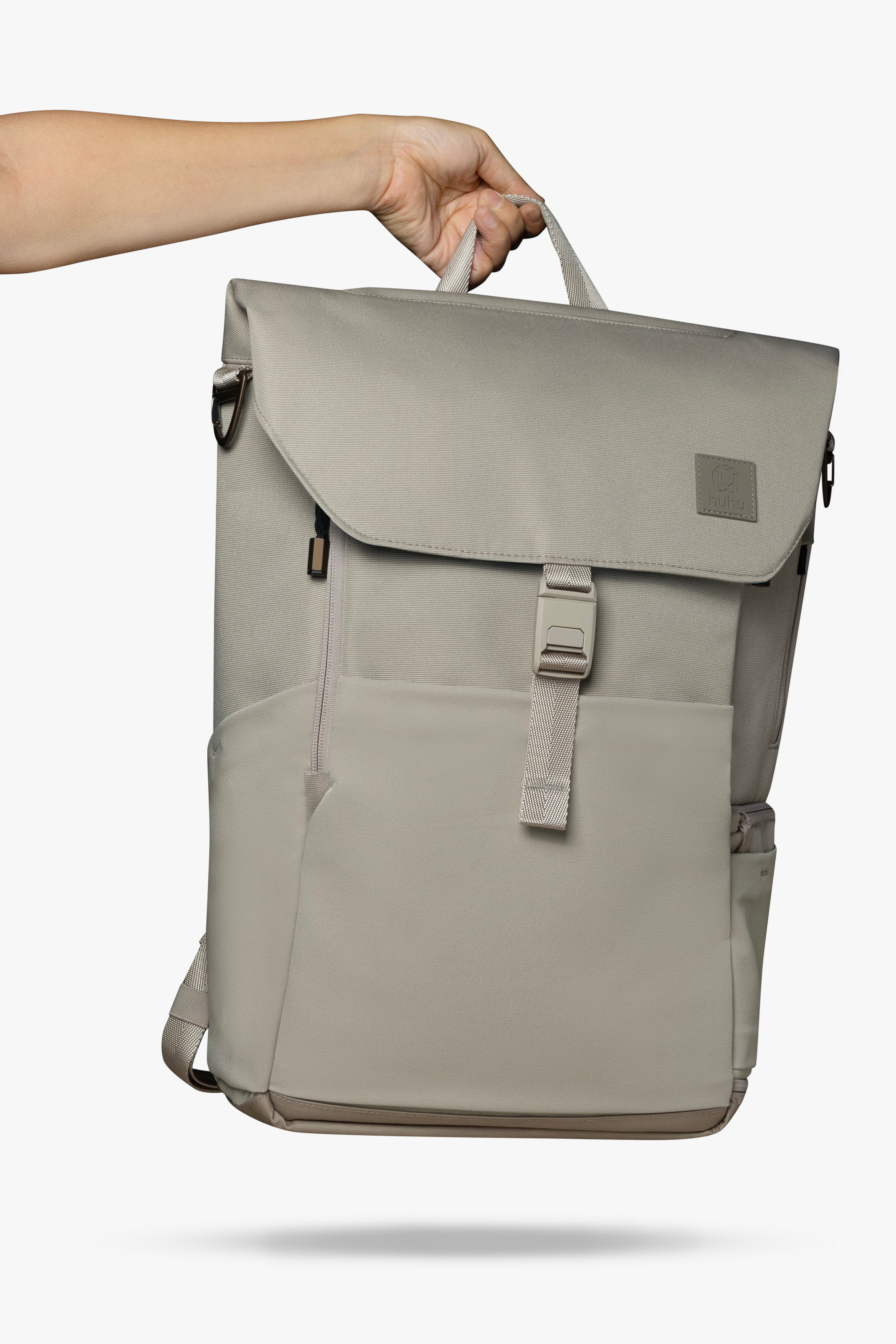 Hand holding taupe backpack by its top handle against white background.