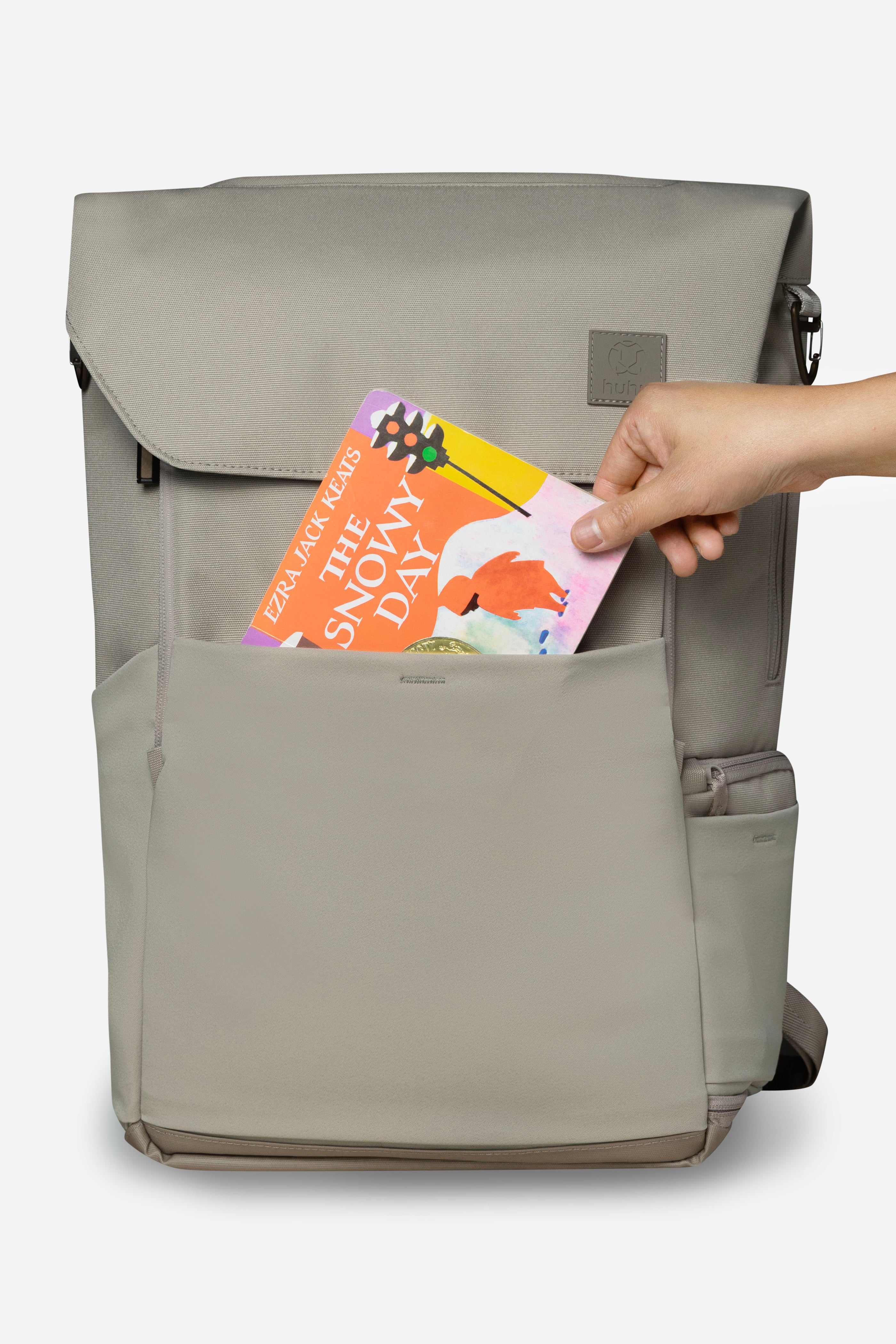 Front view of taupe backpack against white background. Hand is placing orange and yellow board book into front stretchy pocket of backpack.