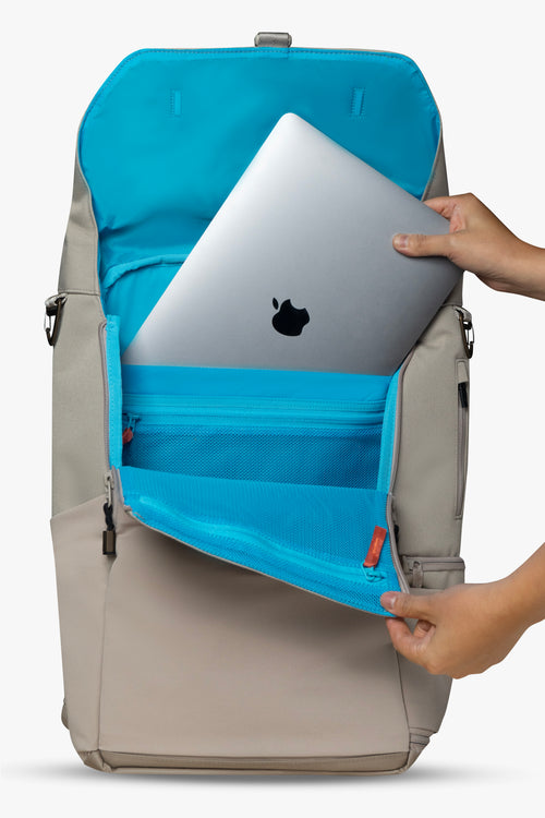 Front view of hands placing silver laptop into padded laptop pocket of an opened taupe backpack against a white background. Interior of bag is blue, with an orange zipper pull showing.
