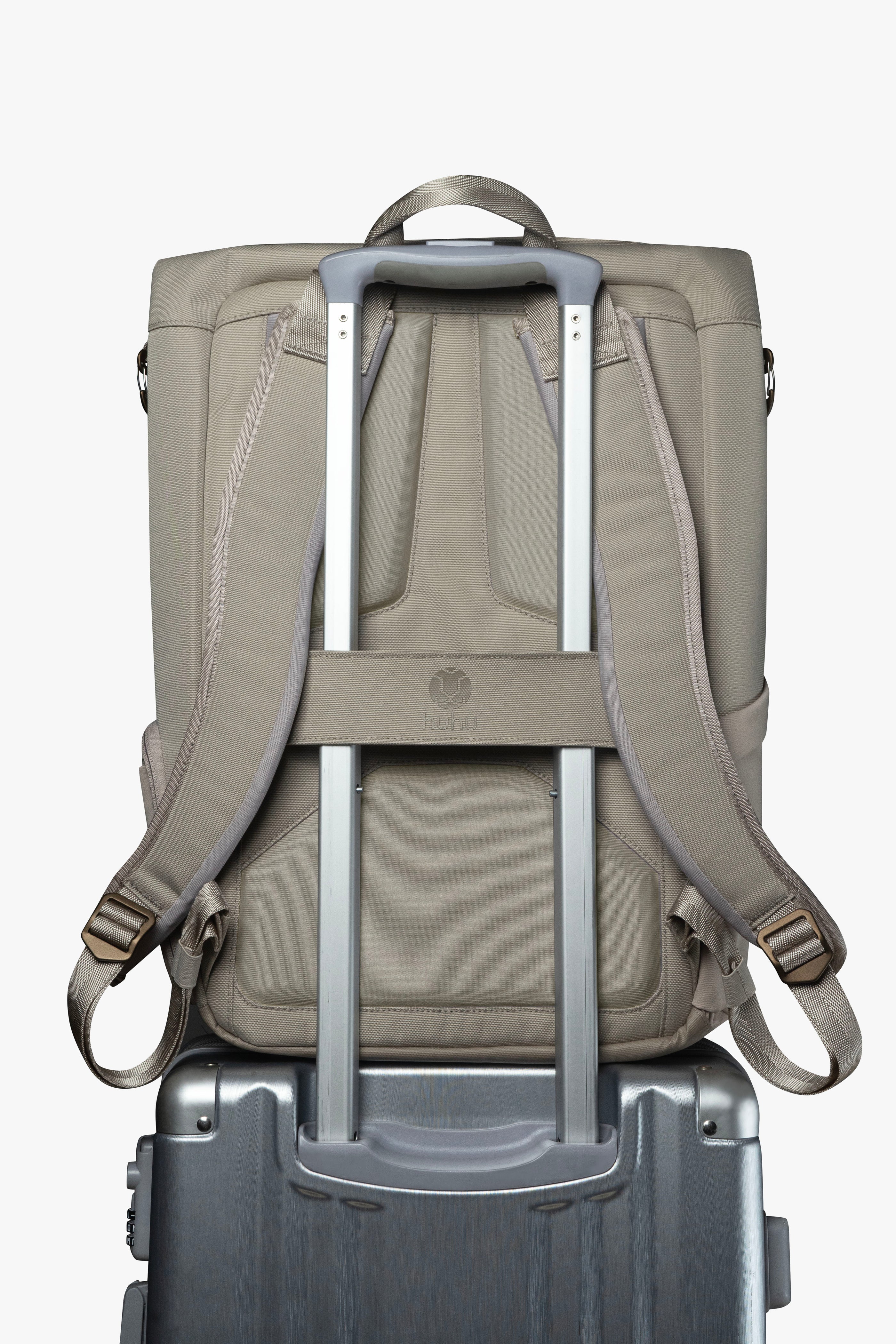 Back view of taupe backpack. Attached luggage strap is holding backpack to the handle of a grey rolling suitcase.