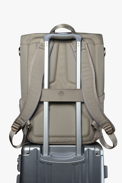 Back view of taupe backpack. Attached luggage strap is holding backpack to the handle of a grey rolling suitcase.
