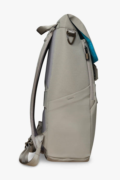 Side view of taupe backpack showing stretchy bottle pocket, padded strap, and bronze carabiner clip against a white background. Blue interior of bag shows on top flap of backpack.