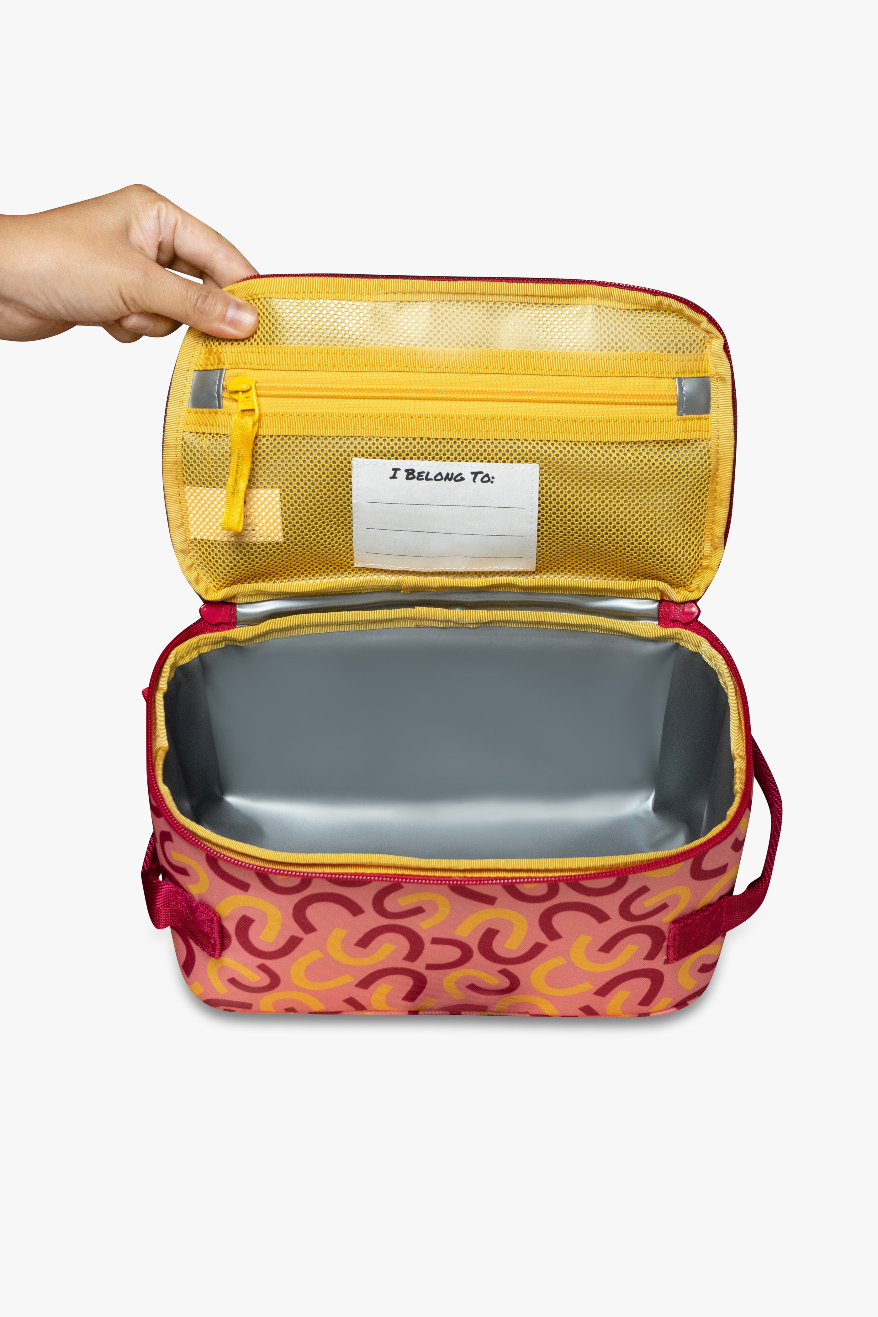 Front view of hand opening pink and yellow macaroni patterned insulated cooler bag to reveal yellow mesh zippered pocket, white "I belong to" tag, and silver interior against a white background.